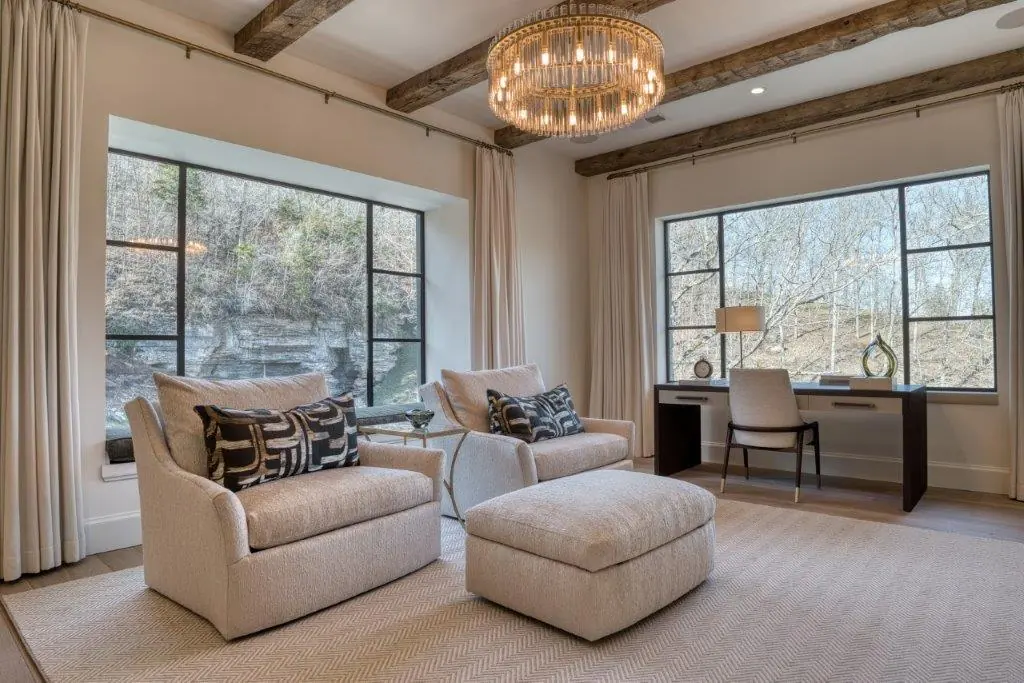 Sitting room with two large black steel windows with divided lights framing a wooded bluff, set beneath reclaimed timber beams