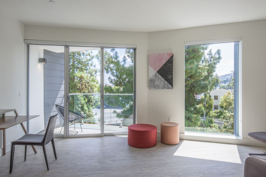 Apartment living room with a full-height aluminum sliding door to a balcony and a large fixed aluminum window framing mature trees, Studio City